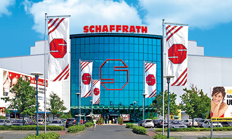 Das Bild zeigt ein modernes Schaffrath-Gebäude mit einer markanten, blau getönten Glasfassade, auf der ein großes, rotes „S“-Logo prangt. Oberhalb der Glasfassade befindet sich der Schriftzug „SCHAFFRATH“ in großen roten Buchstaben. Links und rechts vor dem Gebäude stehen mehrere Fahnenmasten mit weißen Fahnen, die ebenfalls das rote „S“-Logo und rote Streifen zeigen. Im Vordergrund führt ein gepflasterter Weg durch einen gepflegten Grünbereich mit Sträuchern und Bäumen zum Eingang des Gebäudes. Auf der rechten Seite ist ein weiteres Werbeplakat mit einer lächelnden Frau zu sehen. Der Himmel ist blau mit einigen weißen Wolken und verleiht der Szene eine freundliche und einladende Atmosphäre.